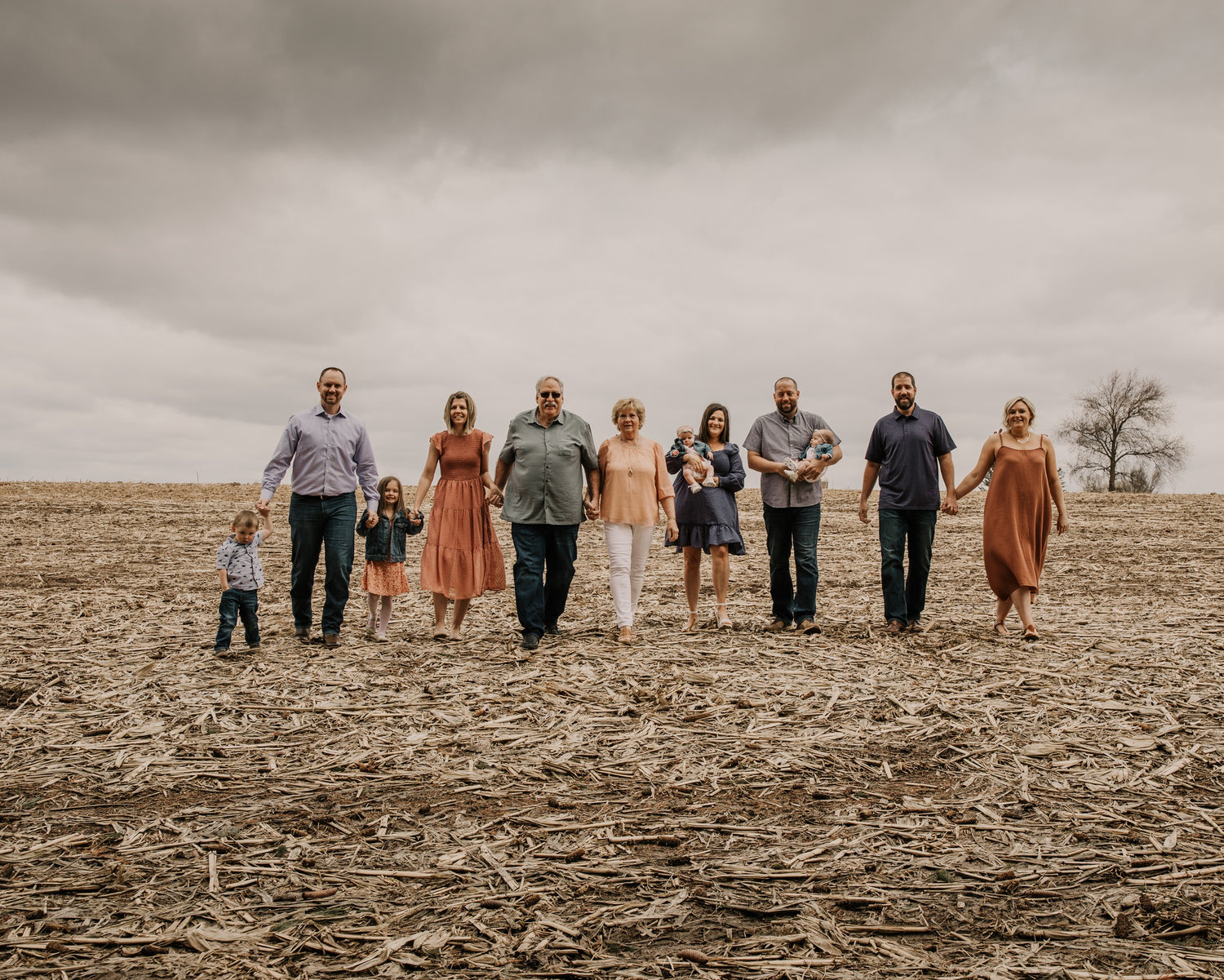 The Knutson family of eight stands together holding hands in a harvested field under a cloudy, overcast sky. The group is dressed in casual, muted-toned clothing, with the ground covered in dry corn stalks.