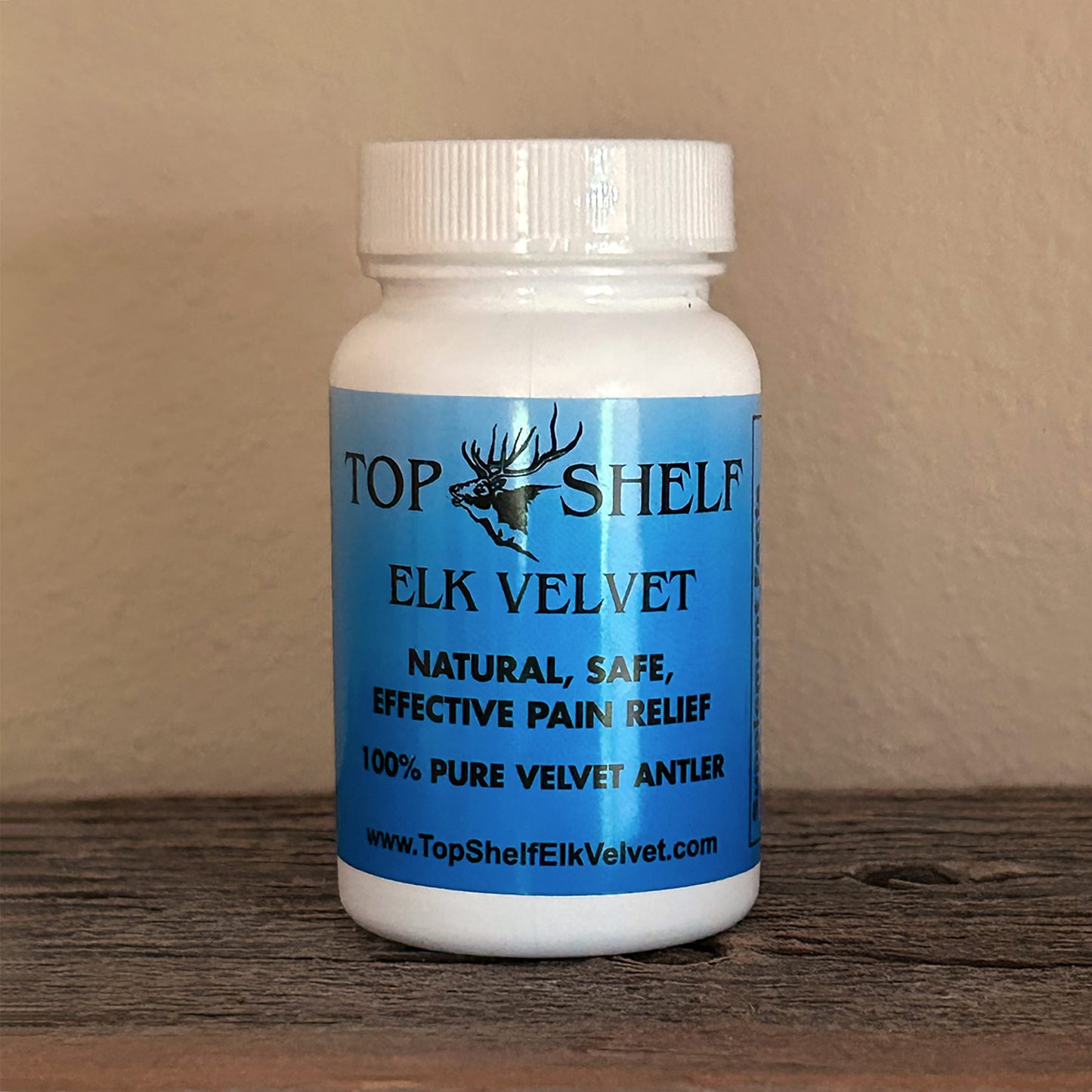 A white plastic bottle with blue label and black letters of Top Shelf Elk Velvet pain relief supplement with website url at the bottom, sitting on a dark wooden surface against a beige wall.
