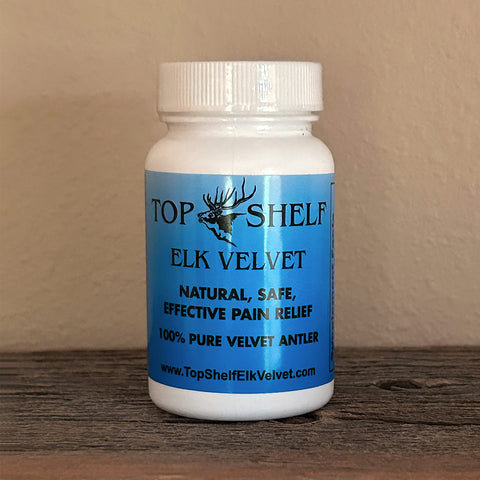 A white plastic bottle of Top Shelf Elk Velvet pain relief supplement with a blue label, sitting on a dark wooden surface against a beige wall.