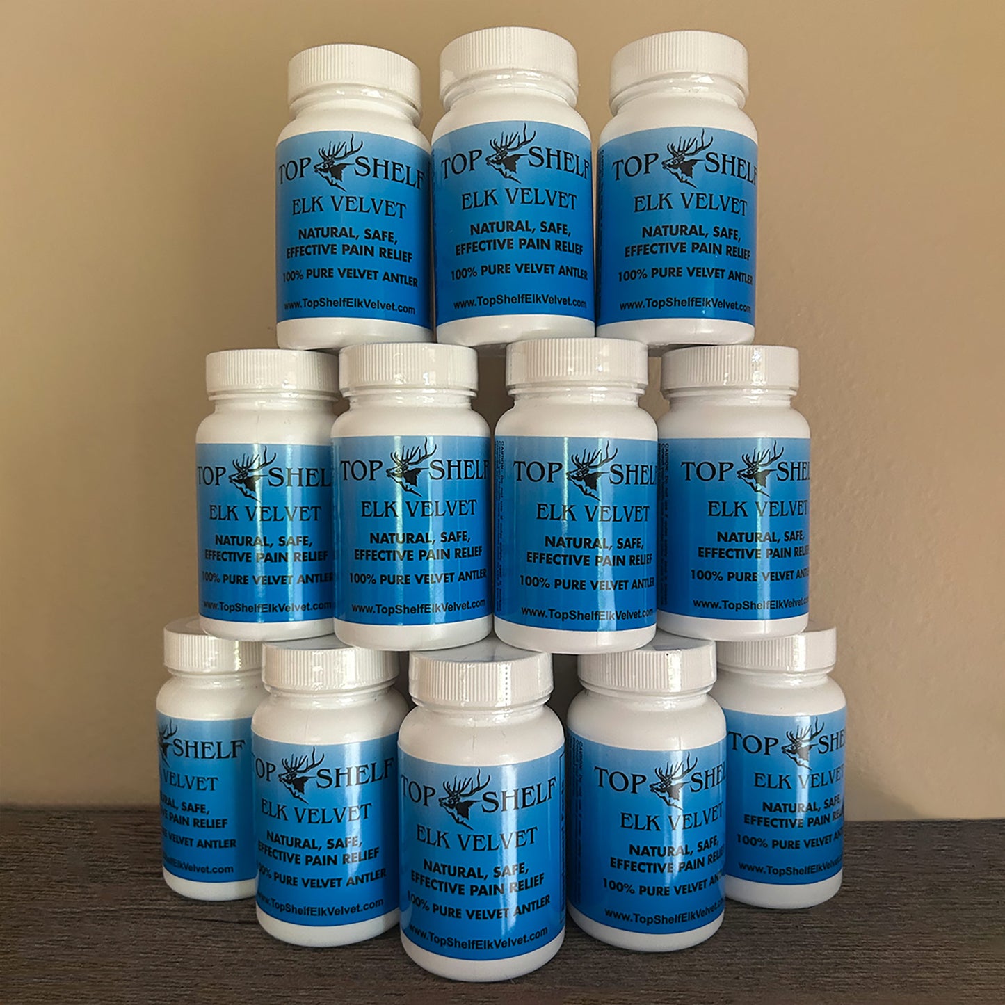 Twelve white plastic supplement bottles with blue labels stacked in three rows on a dark wooden surface against a beige wall. The labels read 'TOP SHELF ELK VELVET NATURAL SAFE EFFECTIVE PAIN RELIEF 100% PURE VELVET ANIMAL'.