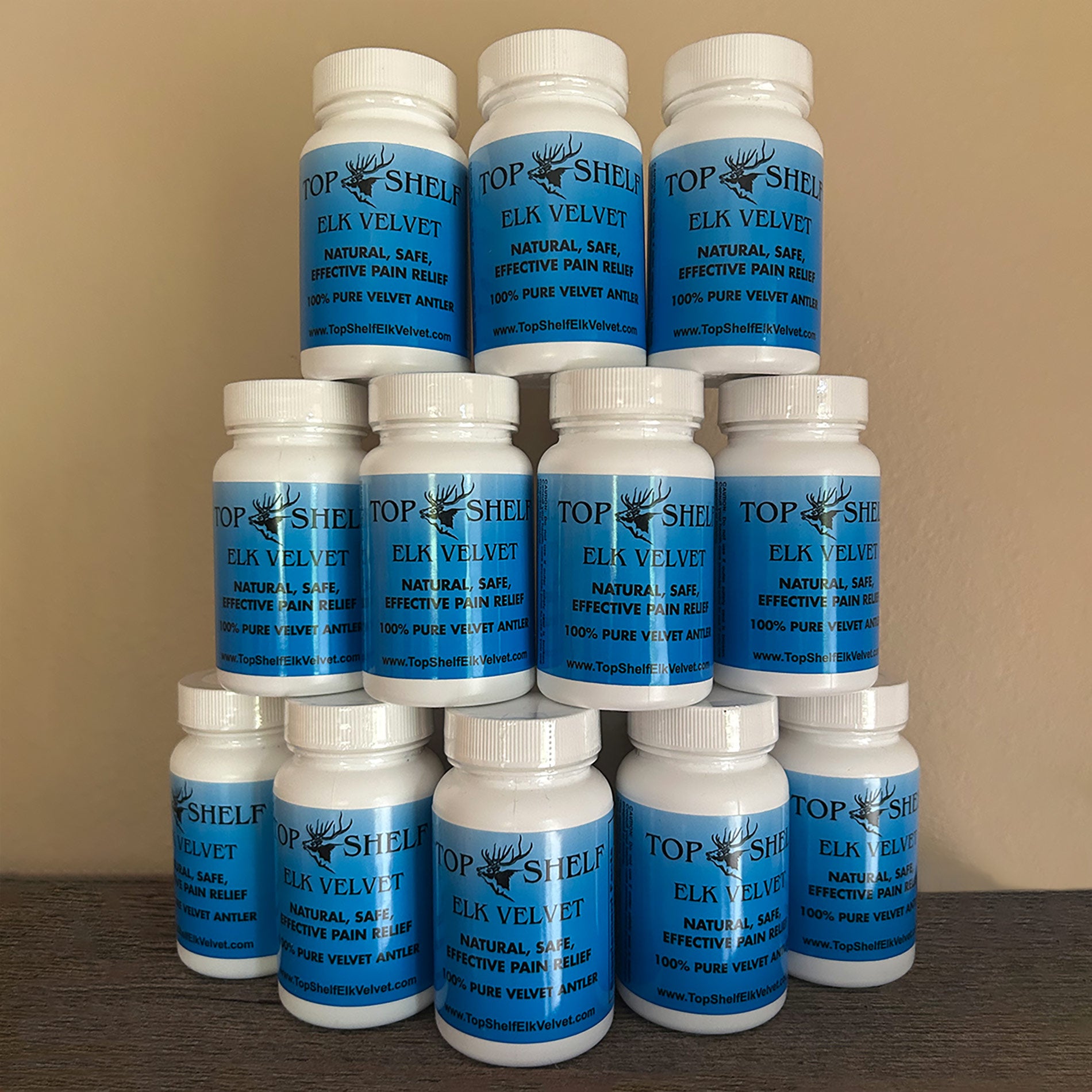 Twelve white plastic supplement bottles with blue labels stacked in three rows on a dark wooden surface against a beige wall. The labels read 'TOP SHELF ELK VELVET NATURAL SAFE EFFECTIVE PAIN RELIEF 100% PURE VELVET ANIMAL'.