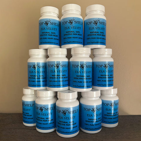 Twelve white plastic supplement bottles with blue labels stacked in three rows on a dark wooden surface against a beige wall. The labels read 'TOP SHELF ELK VELVET NATURAL SAFE EFFECTIVE PAIN RELIEF 100% PURE VELVET ANIMAL'.