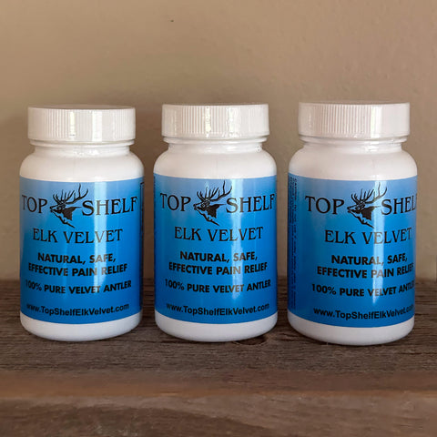 Three white plastic bottles of Top Shelf Elk Velvet natural pain relief capsules lined up on a wooden surface against a beige wall.