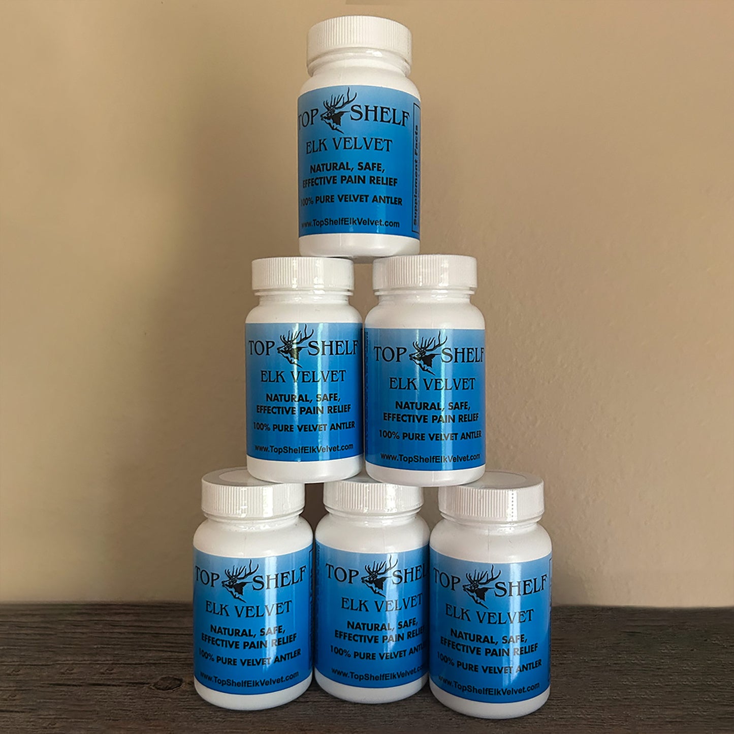 Five white plastic supplement bottles with blue labels stacked in a pyramid arrangement on a dark wooden surface against a beige wall. The labels read 'TOP SHELF ELK VELVET'.