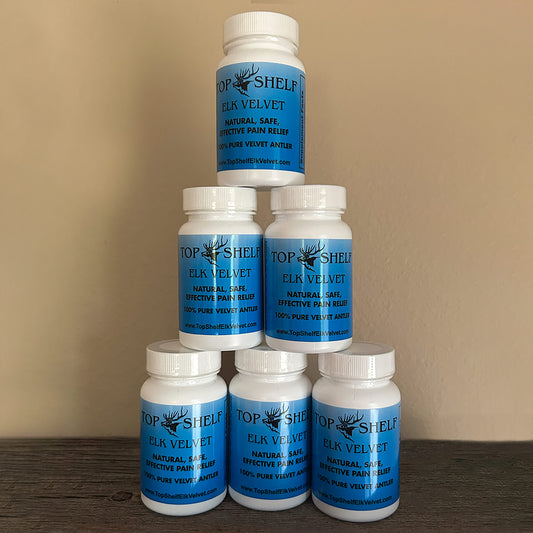 Five white plastic supplement bottles with blue labels stacked in a pyramid arrangement on a dark wooden surface against a beige wall. The labels read 'TOP SHELF ELK VELVET'.