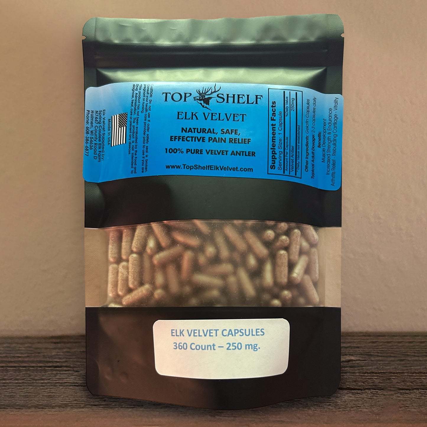 A stand-up pouch of Top Shelf Elk Velvet capsules, showing the beige capsules through a clear window on a wooden surface against a beige wall.
