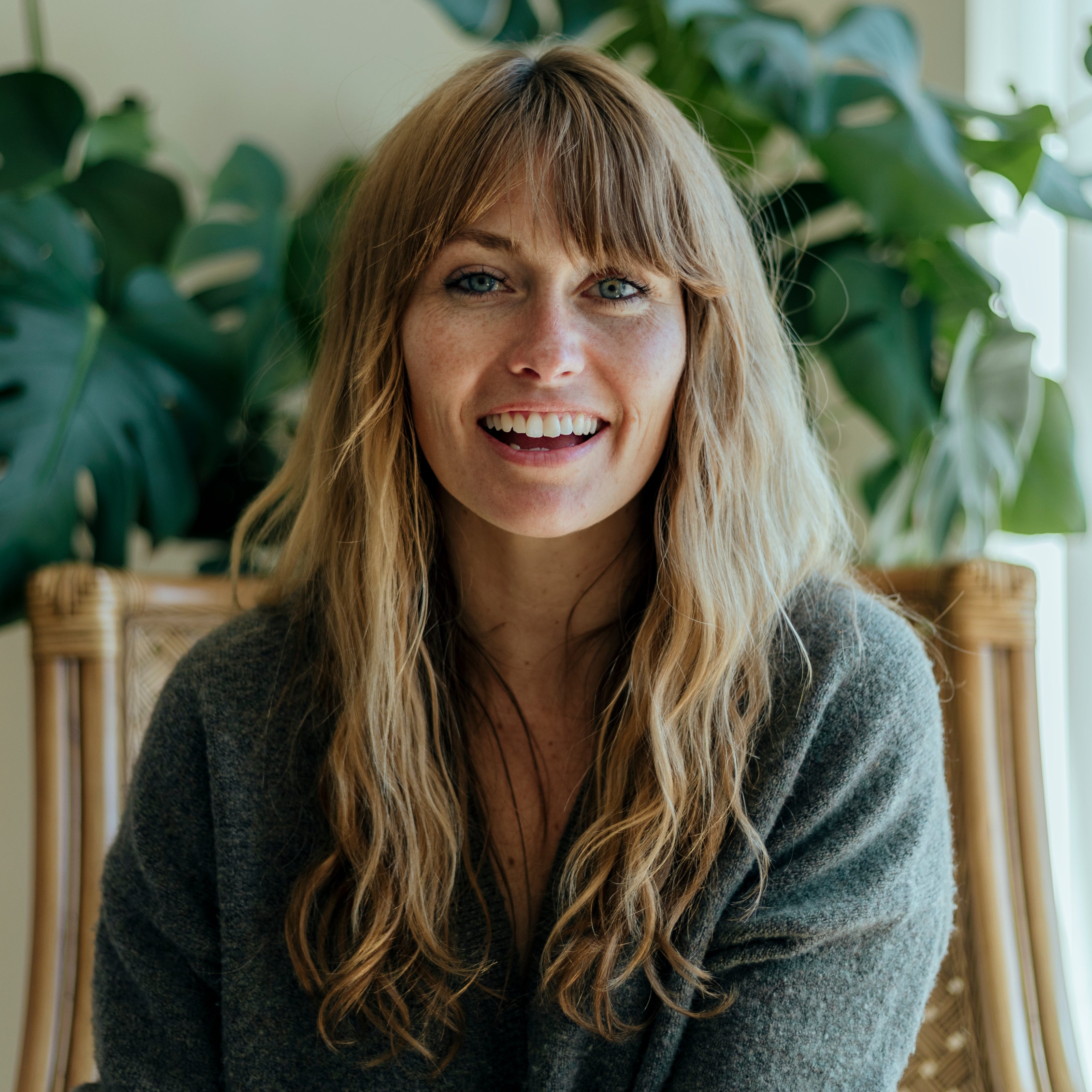 A smiling woman with long blonde hair and bangs, wearing a dark gray knit top, seated in front of large green houseplants.