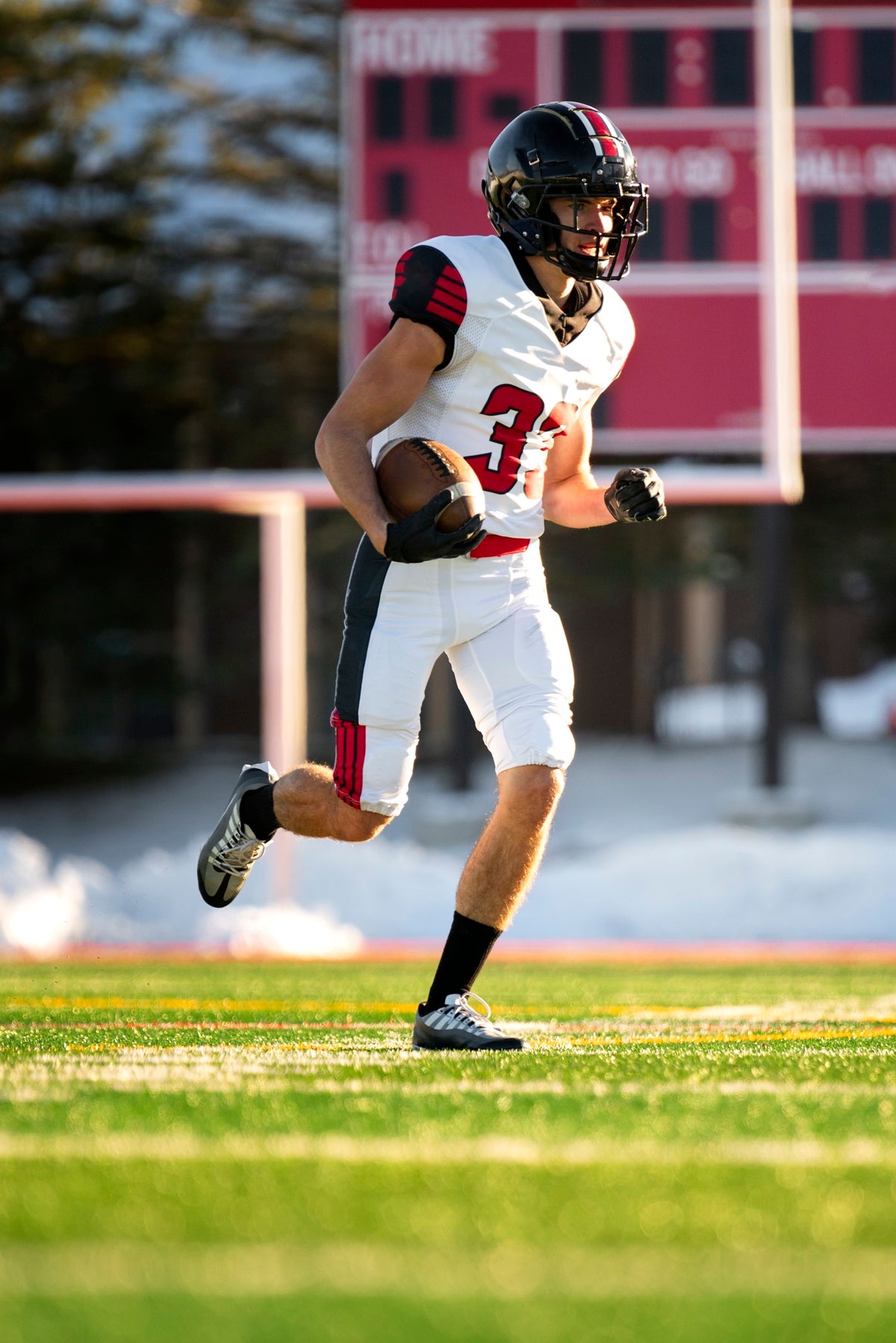 A football player in a white, red, and black uniform runs across a grassy field holding a football, with a blurred scoreboard and trees in the background.