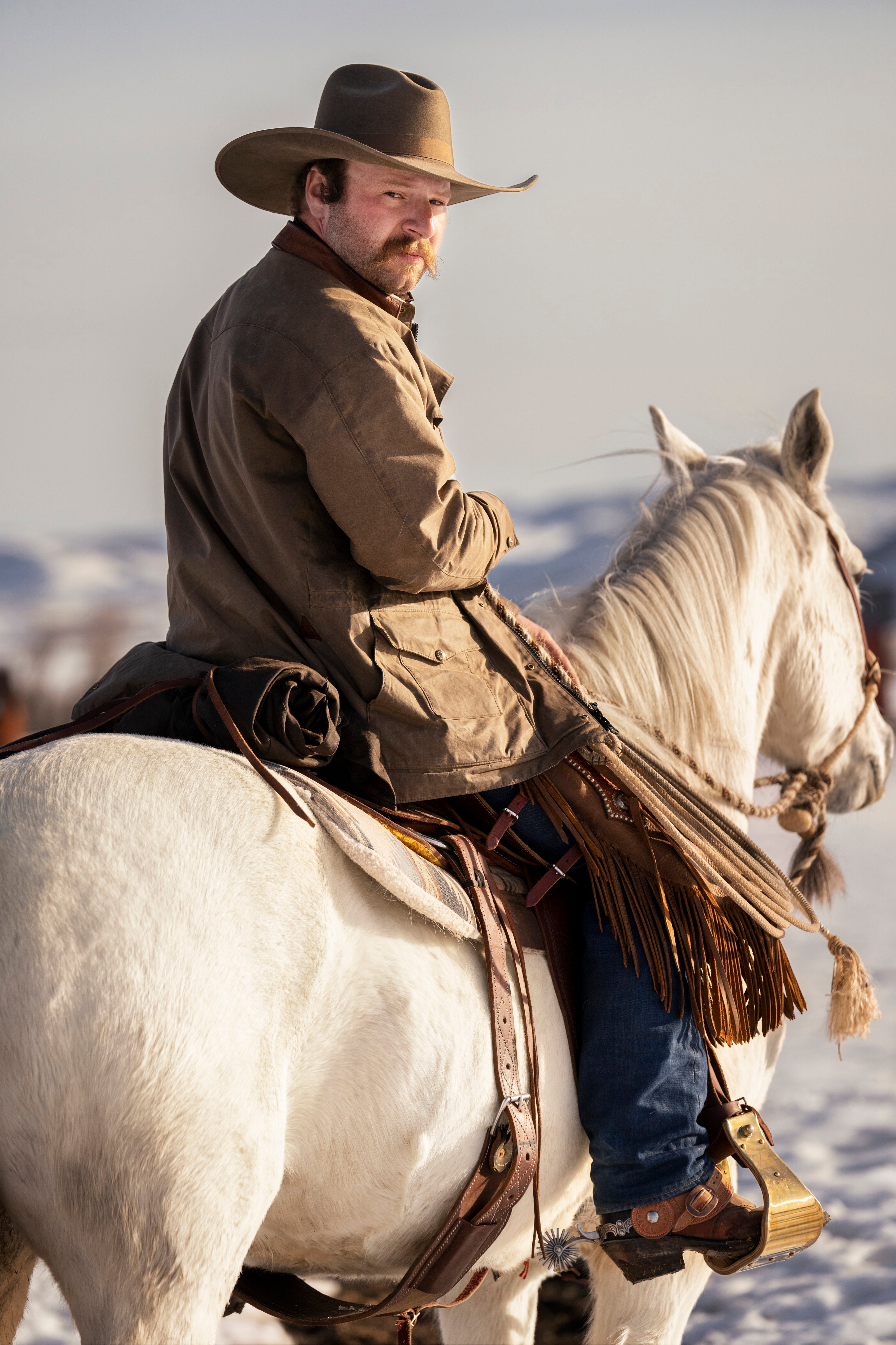 A man wearing a brown cowboy hat, brown jacket, and blue jeans rides a white horse in a snowy outdoor setting.