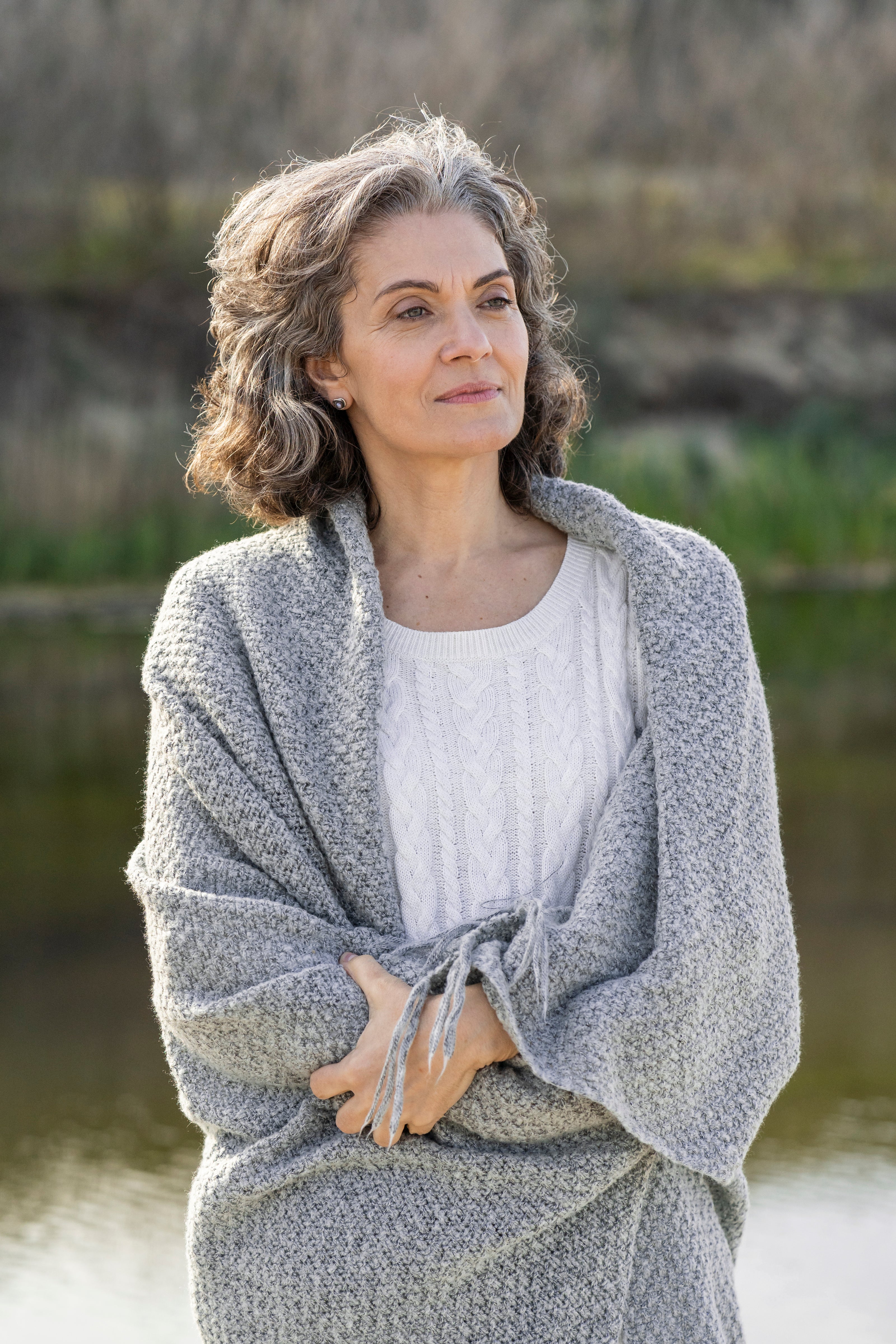 Woman wearing a gray cardigan standing outdoors by a lake